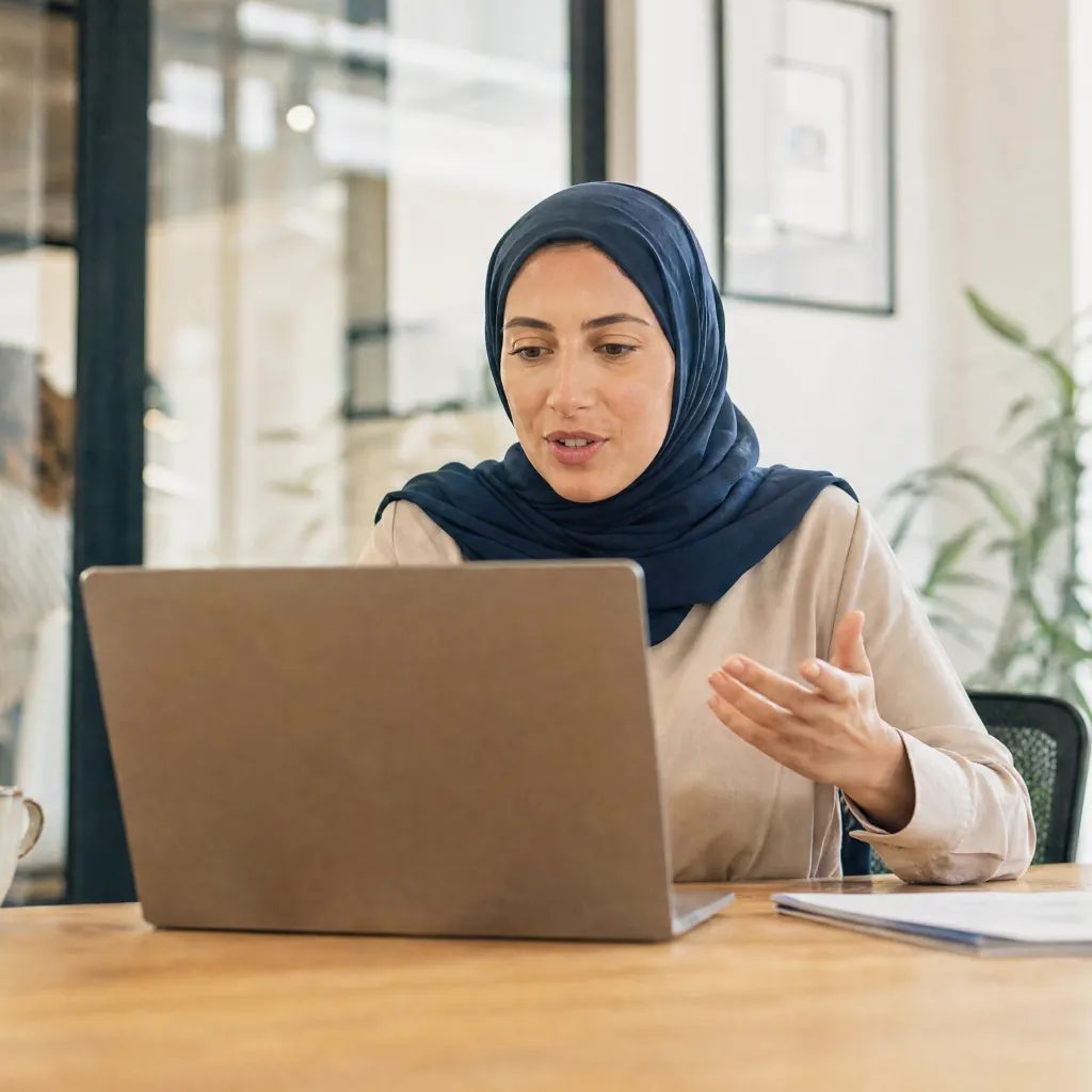 Woman wearing a blue headscarf gesturing toward a laptop at a bright office table.