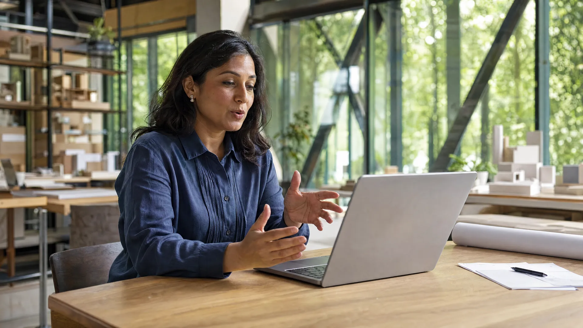 Woman speaking and gesturing beside a laptop at a table in a bright studio.