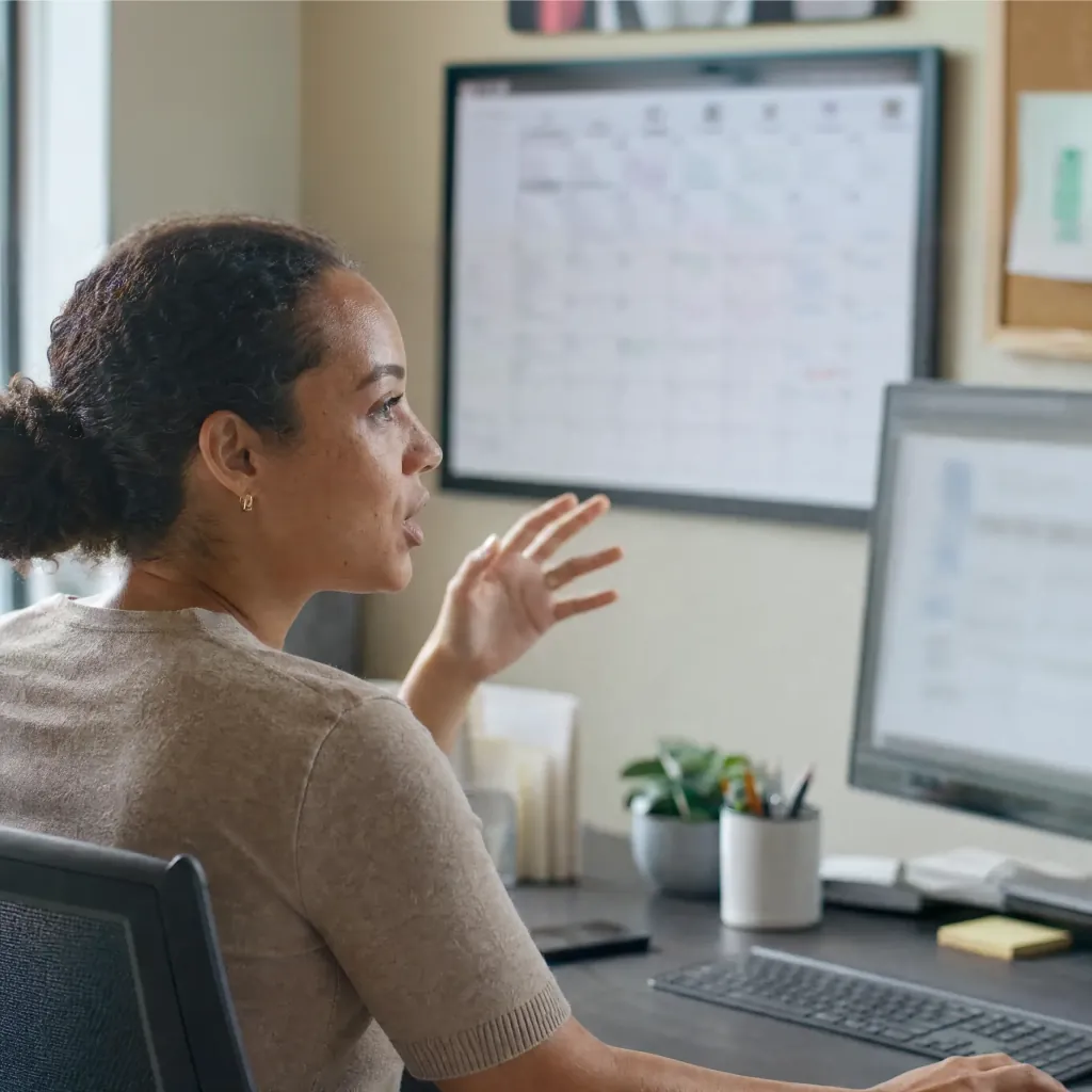 Woman gesturing toward a desktop monitor with a wall calendar behind her