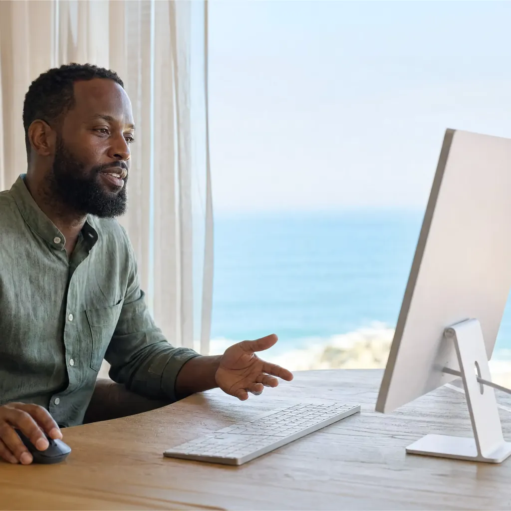 Man speaking toward a desktop monitor at a desk beside a window overlooking the sea.