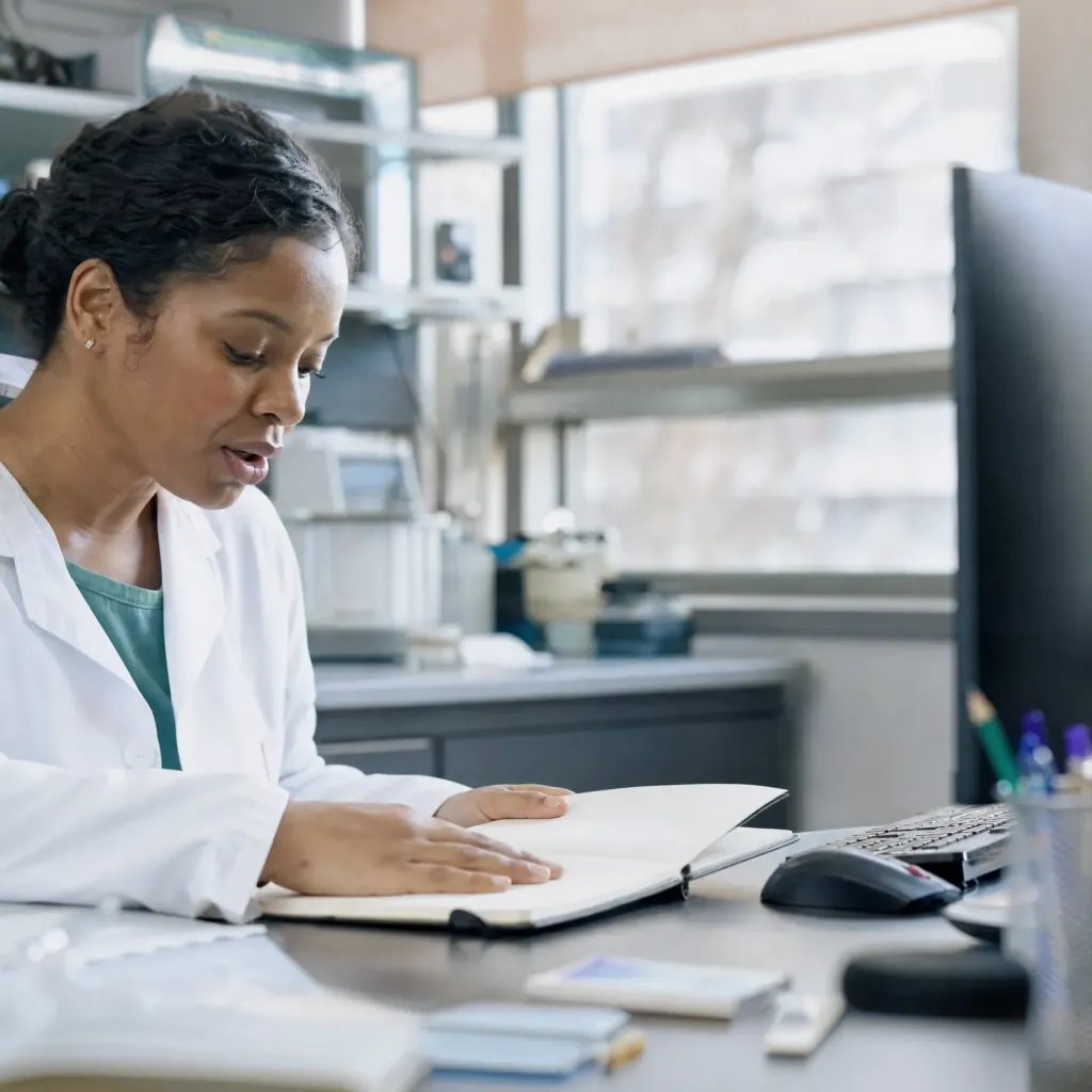 Woman in a white coat looking at an open notebook beside a desktop computer