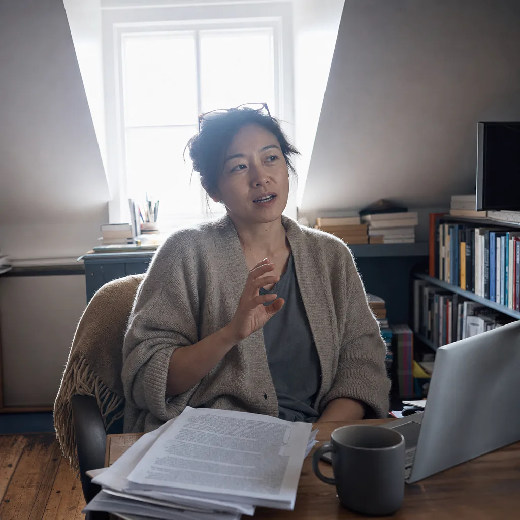 Person speaking at a desk with printed pages and a laptop